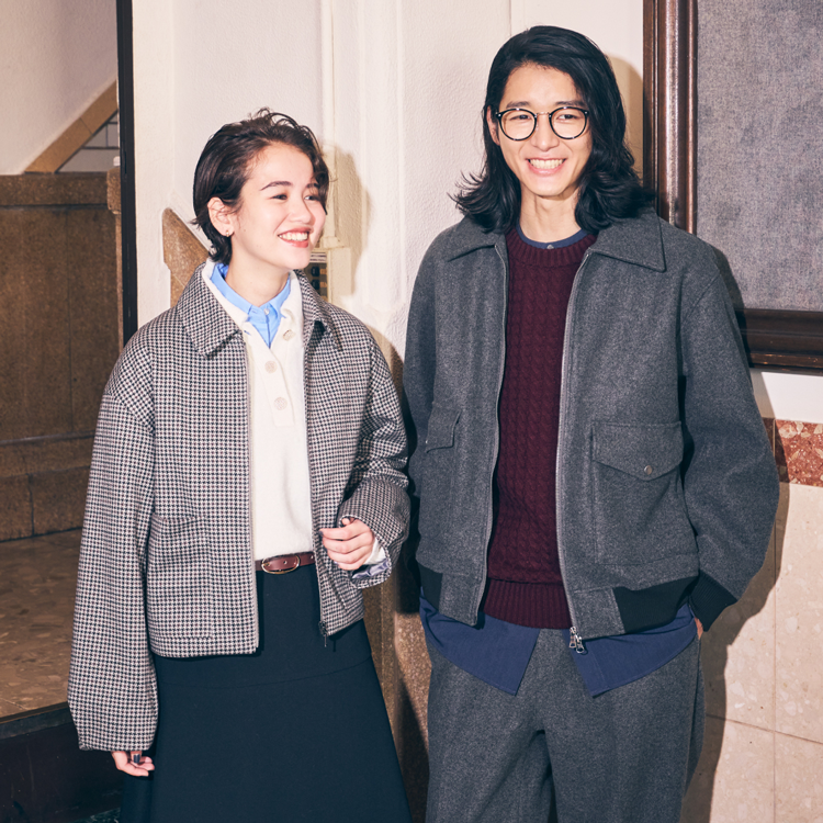 コムサイズム　水色のシャツに白いニット、グレーのブルゾンを羽織り、黒いスカートを履いた女性と、茶色いニットにグレーのブルゾンを羽織り、グレーのズボンを履いた男性の写真